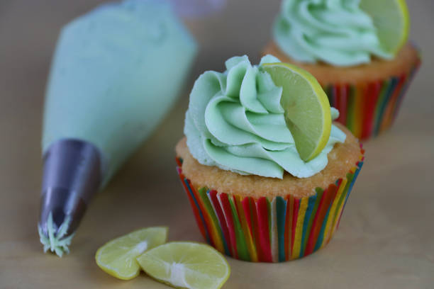 Stock photo showing a batch of pina colada, tropical cocktail cupcakes in paper cake cases sat on sandy beach and clear blue sky background. Afternoon tea concept. imagen de primer plano de lote de cócteles caseros tropicales, cupcakes de piña colada en cajas de pasteles de papel, cubierto con remolinos de glaseado de mantequilla blanca y verde y rodajas de limón, sobre fondo de playa de arena al lado de la bolsa