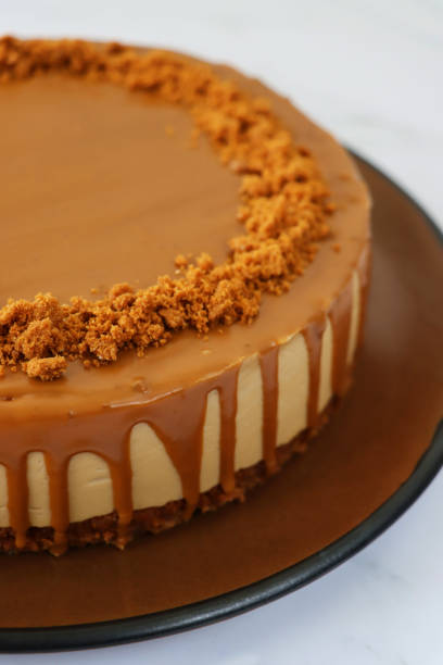 image of uncut toffee caramel cheesecake, buttery biscuit base, fluffy mascarpone cream cheese topping covered with toffee caramel sauce, decorated with biscuit crumbs, served on brown plate against marble effect background - biscuit brown cake unhealthy eating imagens e fotografias de stock