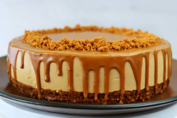 image of whole toffee caramel cheesecake, buttery biscuit base, fluffy mascarpone cream cheese topping covered with toffee caramel sauce, decorated with biscuit crumbs, served on brown plate against marble effect background, focus on foreground - biscuit brown cake unhealthy eating imagens e fotografias de stock