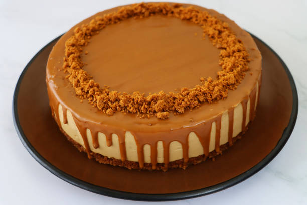 image of whole toffee caramel cheesecake, buttery biscuit base, fluffy mascarpone cream cheese topping covered with toffee caramel sauce, decorated with biscuit crumbs, served on brown plate against marble effect background, elevated view - biscuit brown cake unhealthy eating imagens e fotografias de stock