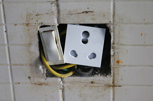 Stock photo showing close-up view of damaged electrical outlet with filthy switch and hole in tiled wall.