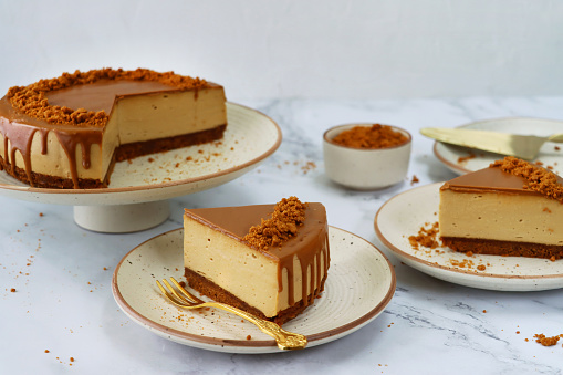 Stock photo showing plates containing sliced toffee caramel cheesecake with buttery biscuit base and fluffy mascarpone cream cheese topping covered with toffee caramel sauce. Served on white plates with brown rims sitting on a marble surface surrounded by gold cutlery and a bowl of crumbed biscuits.