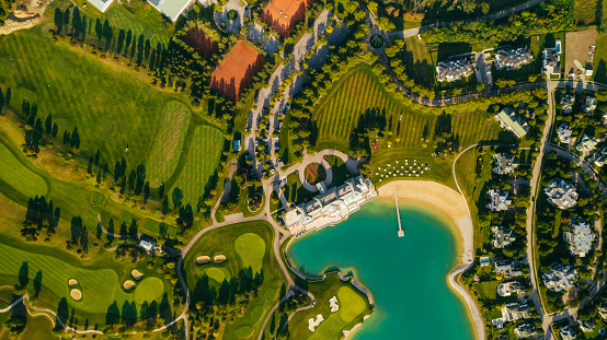 Drone point of view of a golf club near a lake with sand pits, trees, tennis course and a golf mansion. There is also a luxury residential area with modern housing.