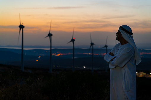 Arab Sheikh with traditional Emirati clothes looking for the energy industry and standing beside a wind turbines farm power station at sunset time