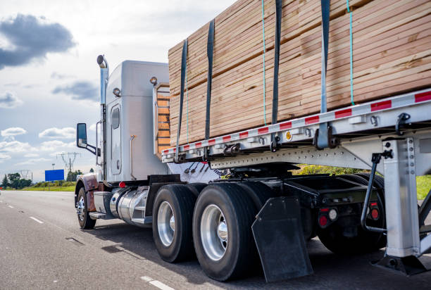 classico trattore semi-camion grande carro che trasporta legname sul semirimorchio a letto piatto che si muove sull'ampia strada autostradale - materiale da costruzione foto e immagini stock