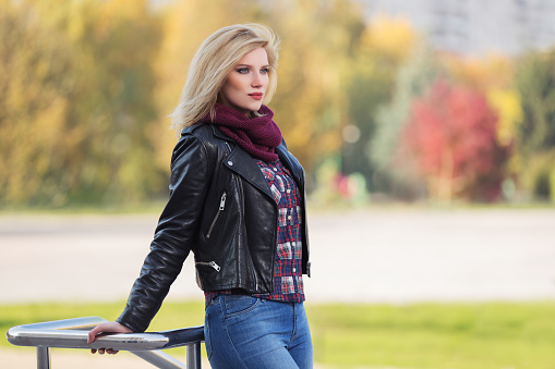 Young fashion blonde woman in city park  Stylish female model in black leather jacket and snood scarf