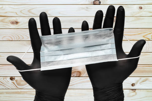 Doctor holds surgical face masks in two hands in black medical gloves on wooden plank brown background. Concept pandemic outbreak, virus prevention stock photo