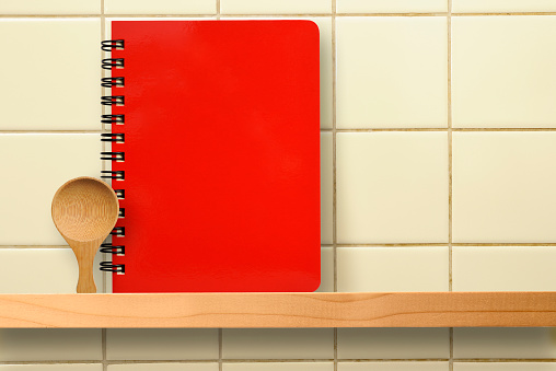 Spiral notebook with wooden spoon on a wooden shelf against old kitchen tiles wall with copy space.