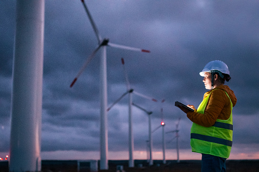 Electrical engineer working for the energy industry, supervising the condition of the Electrical Power Equipment in a wind turbines farm power station at night. Checking the data and the results of measurements with digital tablet. Pregnant woman engineer working on the field. Technology and Global Business.