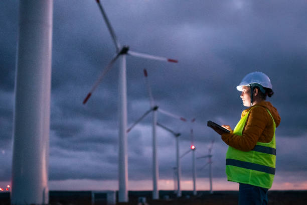Renewable Energy Systems. Electricity Maintenance Engineer working on the field at a Wind Turbine Power station at dusk with a moody sky behind. Blurred motion. Electrical engineer working for the energy industry, supervising the condition of the Electrical Power Equipment in a wind turbines farm power station at night. Checking the data and the results of measurements with digital tablet. Pregnant woman engineer working on the field. Technology and Global Business. industrial windmill stock pictures, royalty-free photos & images