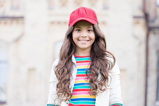 This girl is hipster. Happy little hipster with long curly hair wearing baseball cap outdoor. Cute small hipster smiling with fashionable look. Adorable child in hipster style.