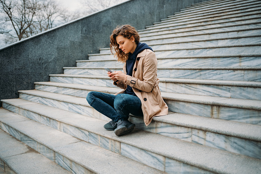 Young woman wearing beige trench coat and blue jeans sitting in city park on gray stone stairs typing in smart phone.