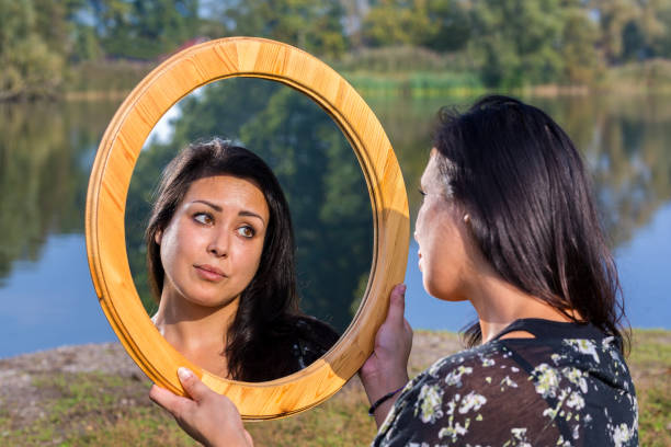 Young moluccan woman looking in round wooden mirror at waterside. The attractive female person is looking at herself on this sunny day in dutch nature.