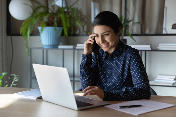 istock Smiling indian woman work on laptop talking on smartphone 1282120162