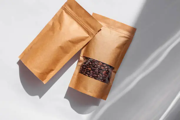 Brown kraft paper pouch bags with coffee beans top view with shadow isolated on white background. Packaging for foods and goods template mockup.Pack with clasp and window for tea leaves weight product