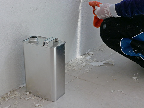 A gallon of thinner placed next to old concrete wall, while a painter is removing sticky rough glue and tape remain on the wall, as a preparation before starting the paint the house