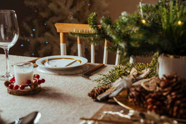 homely christmas table setting, decorated with pine branches and rustic tablecloth in the living room of home tree lit background - christmas table imagens e fotografias de stock