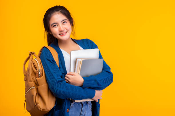 szczęśliwa azjatycka studentka college'u uśmiecha się do kamery na żółtym tle i kopiuje przestrzeń, trzymając laptopa i książki, wieszająca paczkę. młodo-letnia dziewczyna jest studentem wymiany. koncepcja edukacji - education teenagers only studying university zdjęcia i obrazy z banku zdjęć