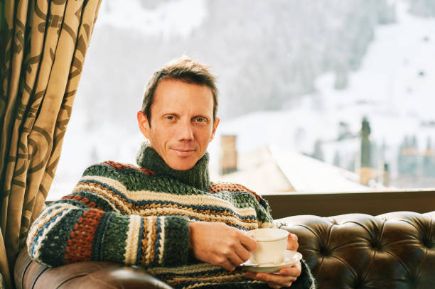 istock Handsome man holding cup of coffee, resting on comfortable leather sofa, ski resort behind the window, relaxing weekend 1282143867
