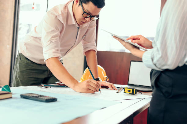 istock engineers holding a pen pointing to a building and  drawing outlay construction plan as guide for builders with details. Engineer design and writing blueprint. 1282150181