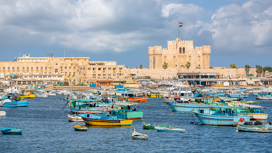 Alexandria, Egypt - November 15, 2018 :  \n\nThe citadel of Qaitbay stands on the site of the ancient lighthouse of Alexandria, one of the seven wonders of the ancient world. Medieval style, this citadel was built to protect the city but it now houses a museum where war remains are exhibited. This place, rich in history, is very popular with tourists and offers a breathtaking view of the Mediterranean and the corniche.\n\nwe see many boats of all sizes facing the citadel. The boats are both pleasure boats and fishing boats passing through or docked. There is really a lot of activity at this marina.\n\nStrategically positioned, 3 sides of the citadel are surrounded by the sea. Near the ramparts, there are a lot of pleasure boats which are are docked in this historic environment.