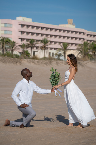 Just married honeymoon wedding couple giving flower bouket on his knees mixed ethnicity on the beach sand happy together