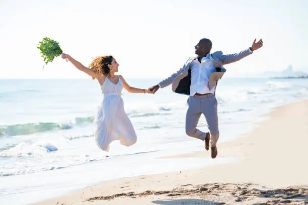 Just married honeymoon wedding couple jumping high happy mixed ethnicity on the beach sand happy together