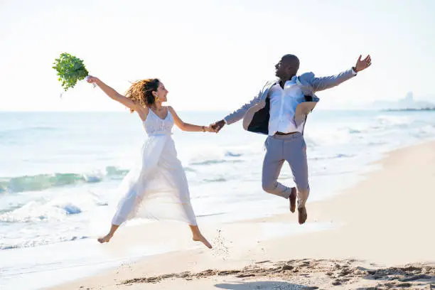 Just married honeymoon wedding couple jumping high happy mixed ethnicity on the beach sand happy together