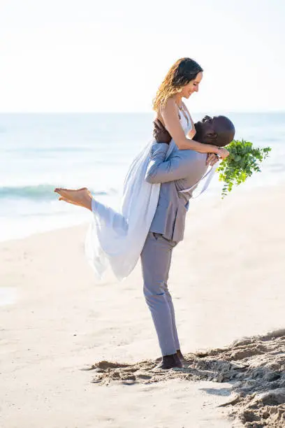 Just married honeymoon wedding couple jumping hug mixed ethnicity on the beach sand happy together