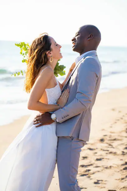 Just married honeymoon wedding couple hug laughing mixed ethnicity on the beach sand happy together