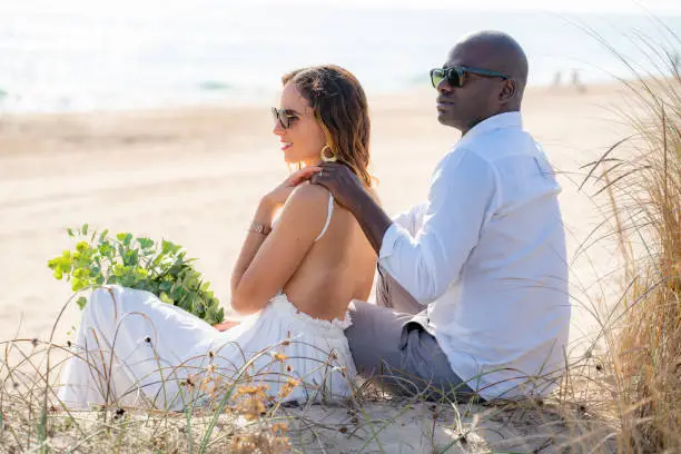 Just married honeymoon wedding couple mixed ethnicity sitting on the beach sand happy together