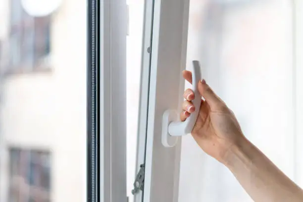 Cropped view of woman holding white modern handle at pvc window with double glazing, opening plastic frame for ventilation her house