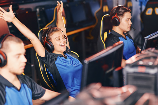 Young excited mixed race girl, female cybersport gamer wearing headphones raising hands up, celebrating success while participating in eSport tournament, playing online video games professionally
