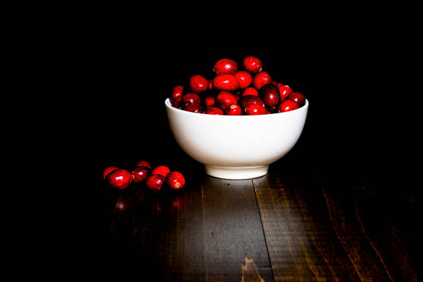 A bunch of cranberries in a white porcelain bowl, with black background and brown wood base. Shot in low key style.