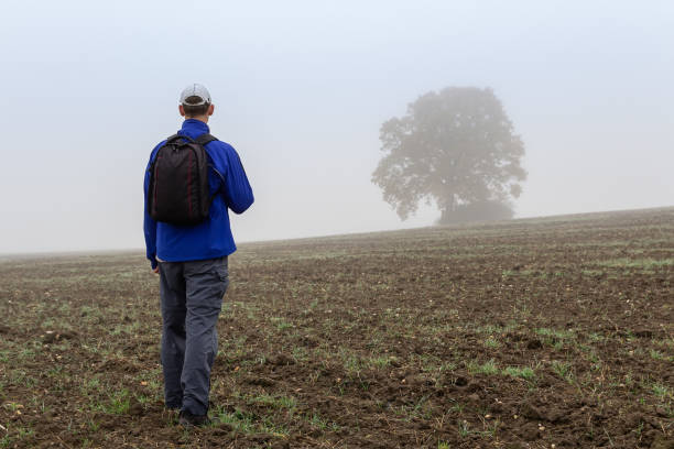istock Young man in blue jacket and white cap with backpack walking on fileld with big tree on autumn misty fog. Czech landscape 1282174989