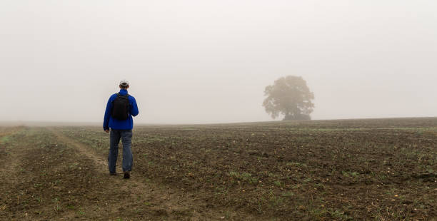 istock Young man in blue jacket and white cap with backpack walking on fileld with big tree on autumn misty fog. Czech landscape 1282175060