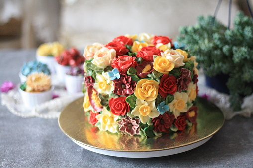 A rainbow flower overlay buttercream flower cake stand on golden pad with several cupcakes. PS: Flowers on the cake are not real, made it with buttercream. The cake made and shot by Seyma Fidan