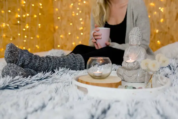 Woman is holding a cup and sitting in the background. In the foreground is a buddha and candles. Fairy lights in the background