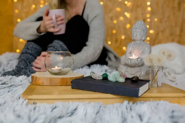 Woman is holding a cup and sitting in the background. In the foreground is a buddha, crystals and a book. Fairy lights in the background
