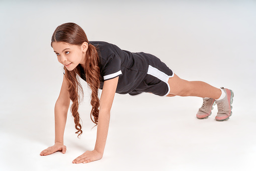 Being physically active. Full length of cute and happy teenage girl in sportswear standing in plank position and smiling isolated over grey background in studio. Kids and sport, healthy living
