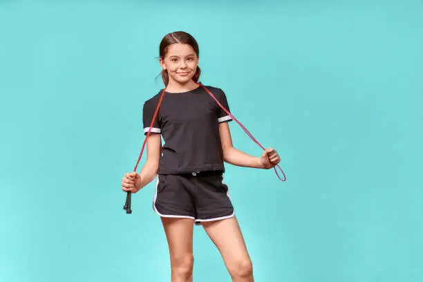 Photo of Cute teenage girl in black sportswear with jumping rope looking at camera and smiling while having workout, standing isolated over blue background