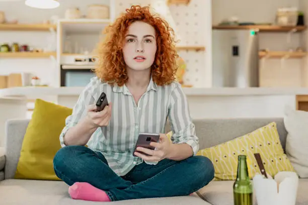 A young woman is watching tv at home, she is sitting on the couch and holding a mobile phone