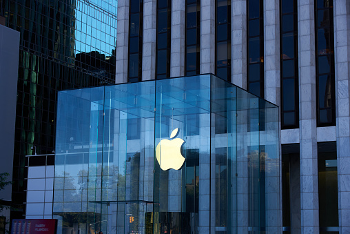 New York, NY - October 14, 2020: The Apple logo is illuminated with a warm light on a dark day at the Apple Store on 5th Avenue, Manhattan. The glass structure of the entry shows the cool blue light on the buildings