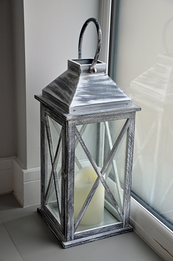Large grey / silver coach lantern with candle on tiled floor near a plain grey wall and frosted window. Domestic entrance hall.