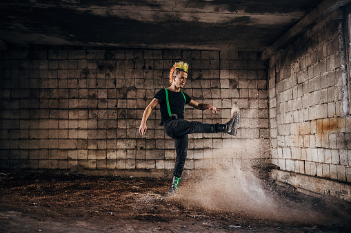 One angry young male punker with colorful coiffure standing in abandoned warehouse and kicking the dust.