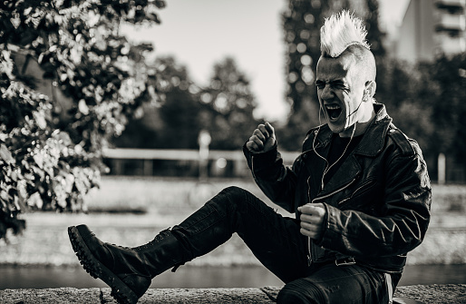 Male punk person with yellow and red Mohawk hairstyle, listening music on headphones and shouting, sitting by the river in city.