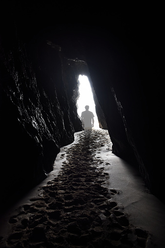 A transparent ghostly figure walks towards the light at the end of a tunnel, heading out of a cave with sandy footprints leading out from the foreground.