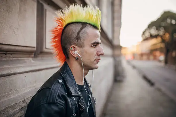 One young male punker with colorful coiffure standing on the city street and listening music on the smart phone.