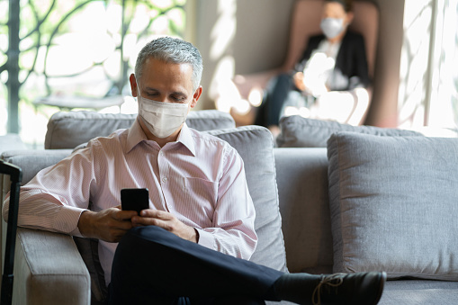 Latin american mature business man texting on smartphone while sitting at hotel lounge wearing a protective face mask during COVID-19