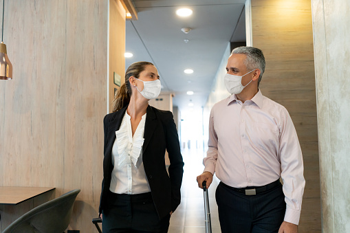 Cheerful business couple looking at each other while leaving hotel both wearing protective face masks - Pandemic Lifestyles